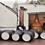 Several wooden barrels are lined up on the ground outside a distillery building, with copper stills visible inside and outside the facility.