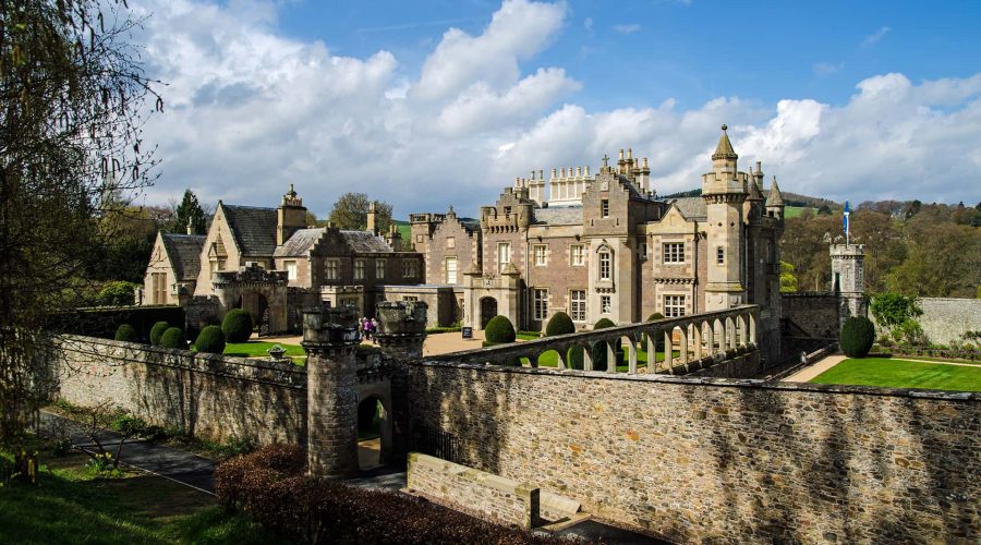 A large stone castle with multiple turrets and arched windows, surrounded by a stone wall and manicured lawns under a partly cloudy sky.