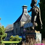 Statue of a piper in traditional dress in front of a historic stone building surrounded by trees and lush greenery under a clear blue sky.