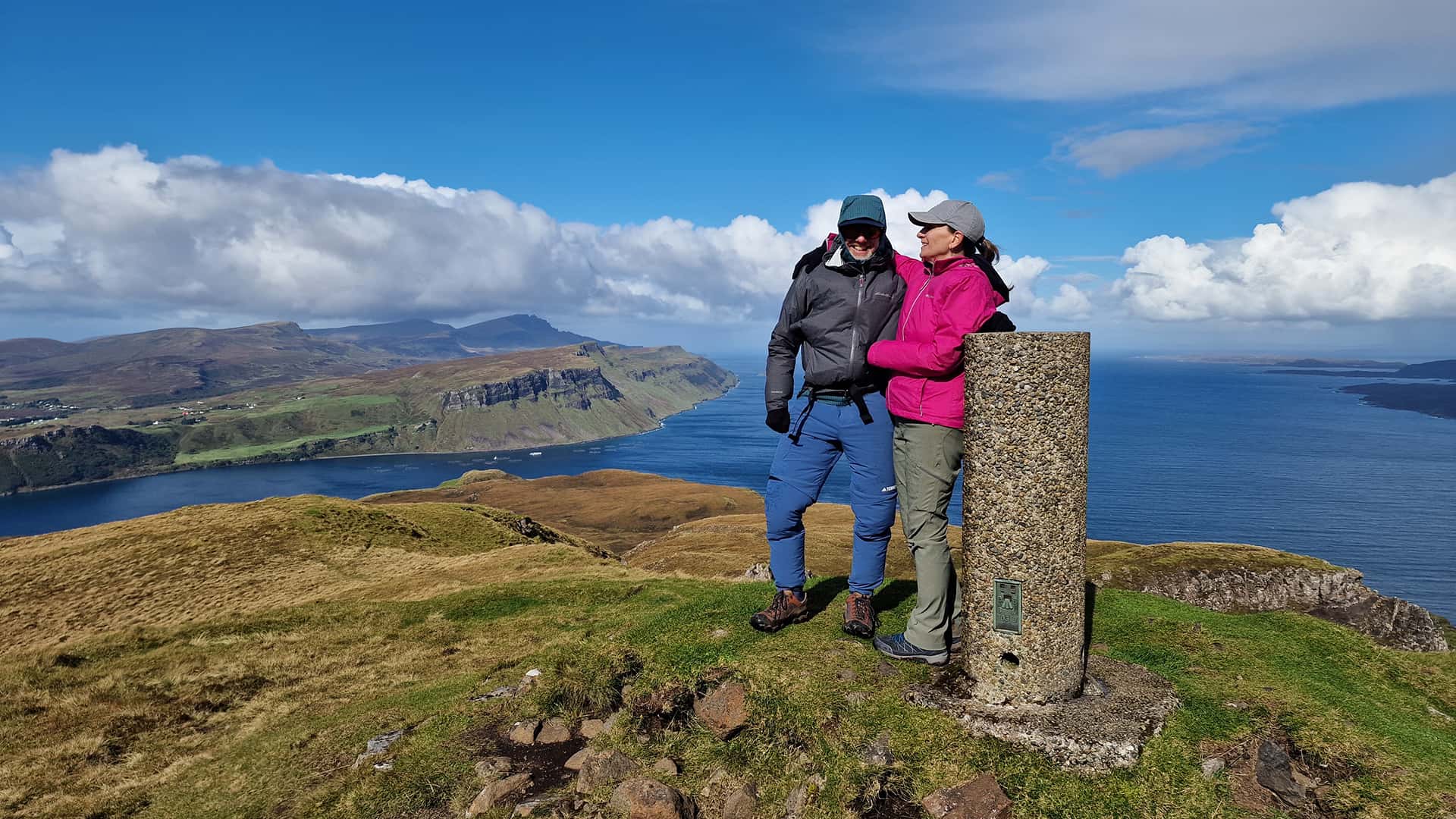 A couple stands on a hilltop near a stone marker, overlooking a scenic coastal landscape with cliffs and open water under a partly cloudy sky.