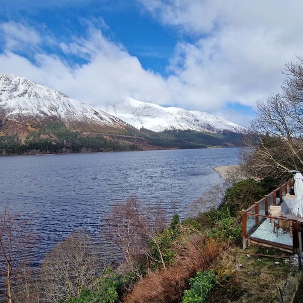 Snow-capped mountains and a lake with a wooden deck on the right, featuring tables and chairs under a partly cloudy sky.