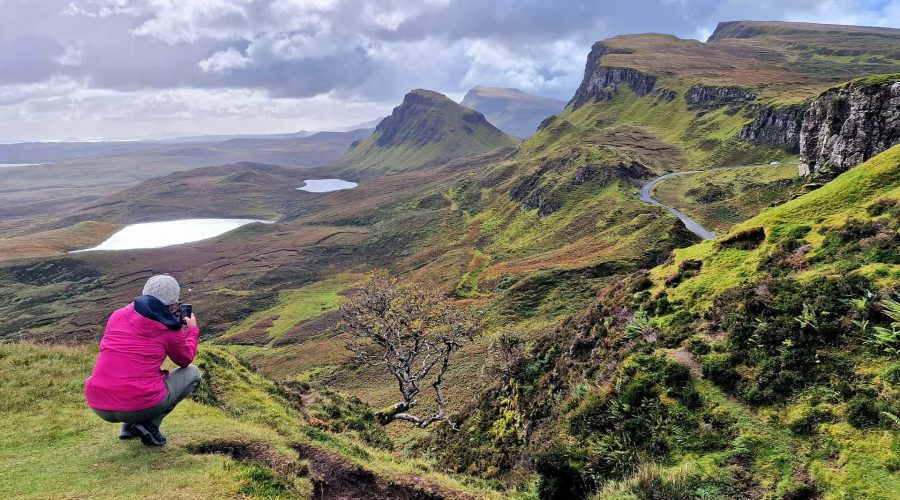 Person in a pink jacket photographs a scenic landscape with lush green hills, a winding road, and small lakes under a cloudy sky.