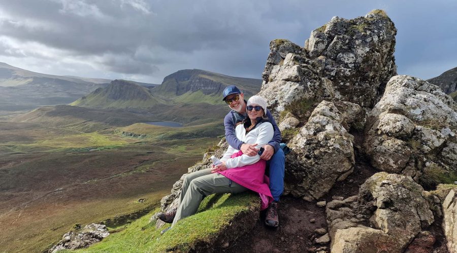 A couple sits on a rocky outcrop with expansive green hills and mountains in the background under a cloudy sky.