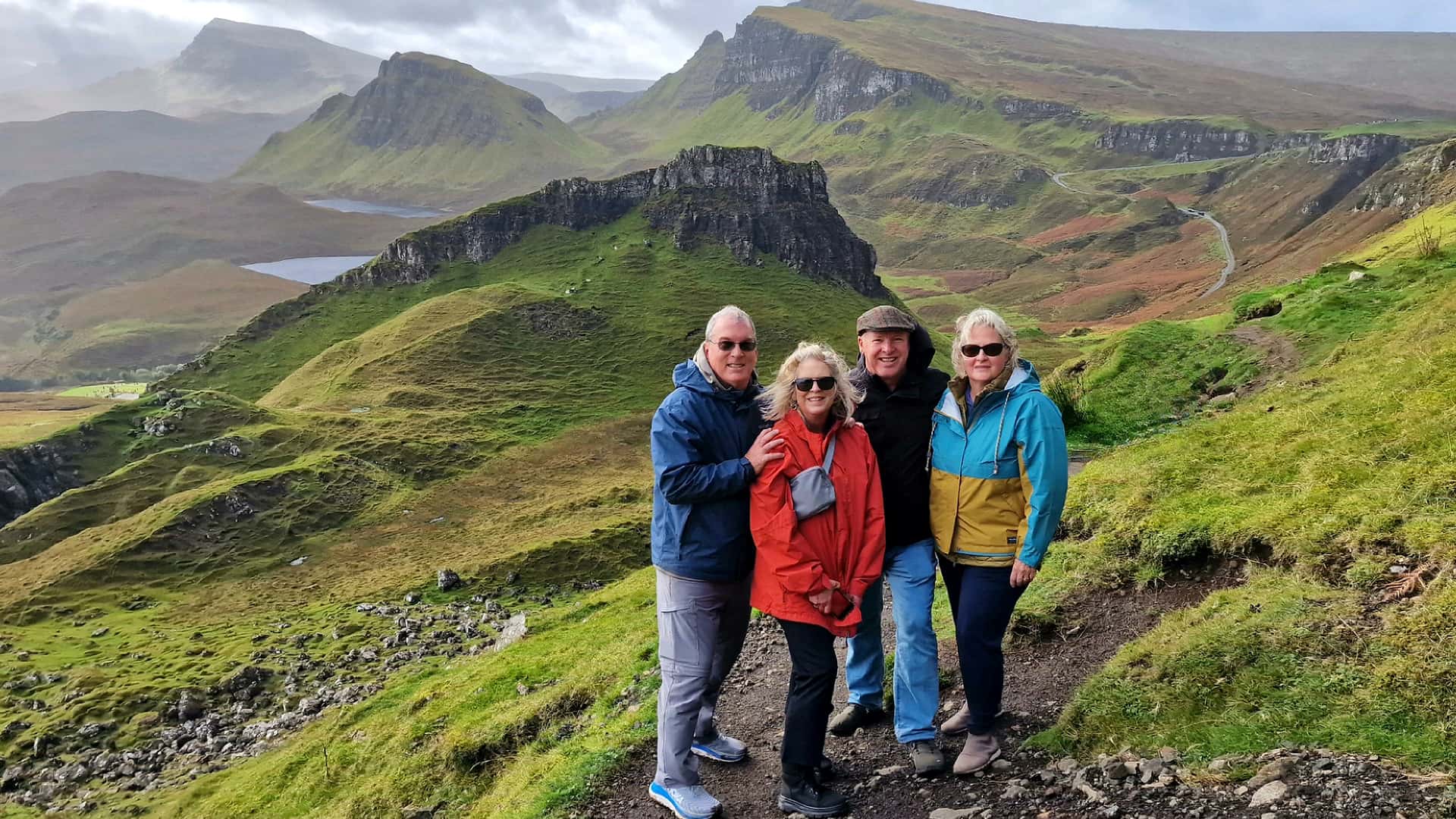 Four people stand on a grassy hill with a backdrop of rugged mountains and a cloudy sky.