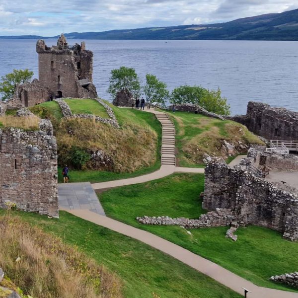 Ruins of Urquhart Castle sit by the edge of Loch Ness, surrounded by grassy areas and pathways, under a cloudy sky.