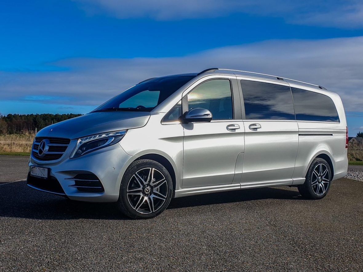 Silver Mercedes-Benz van parked on a paved surface with a backdrop of grass, trees, and a partly cloudy sky. Silver Mercedes-Benz van parked on a paved surface with a backdrop of grass, trees, and a partly cloudy sky.