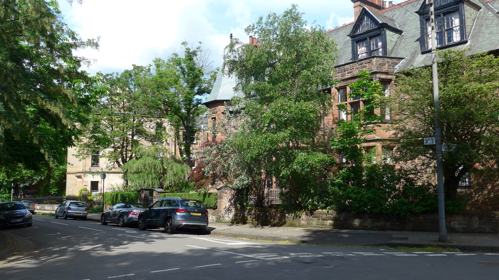 Tree-lined street in Glasgow with parked cars beside large, historic brick buildings partially obscured by greenery on a sunny day—reminiscent of iconic Outlander locations.